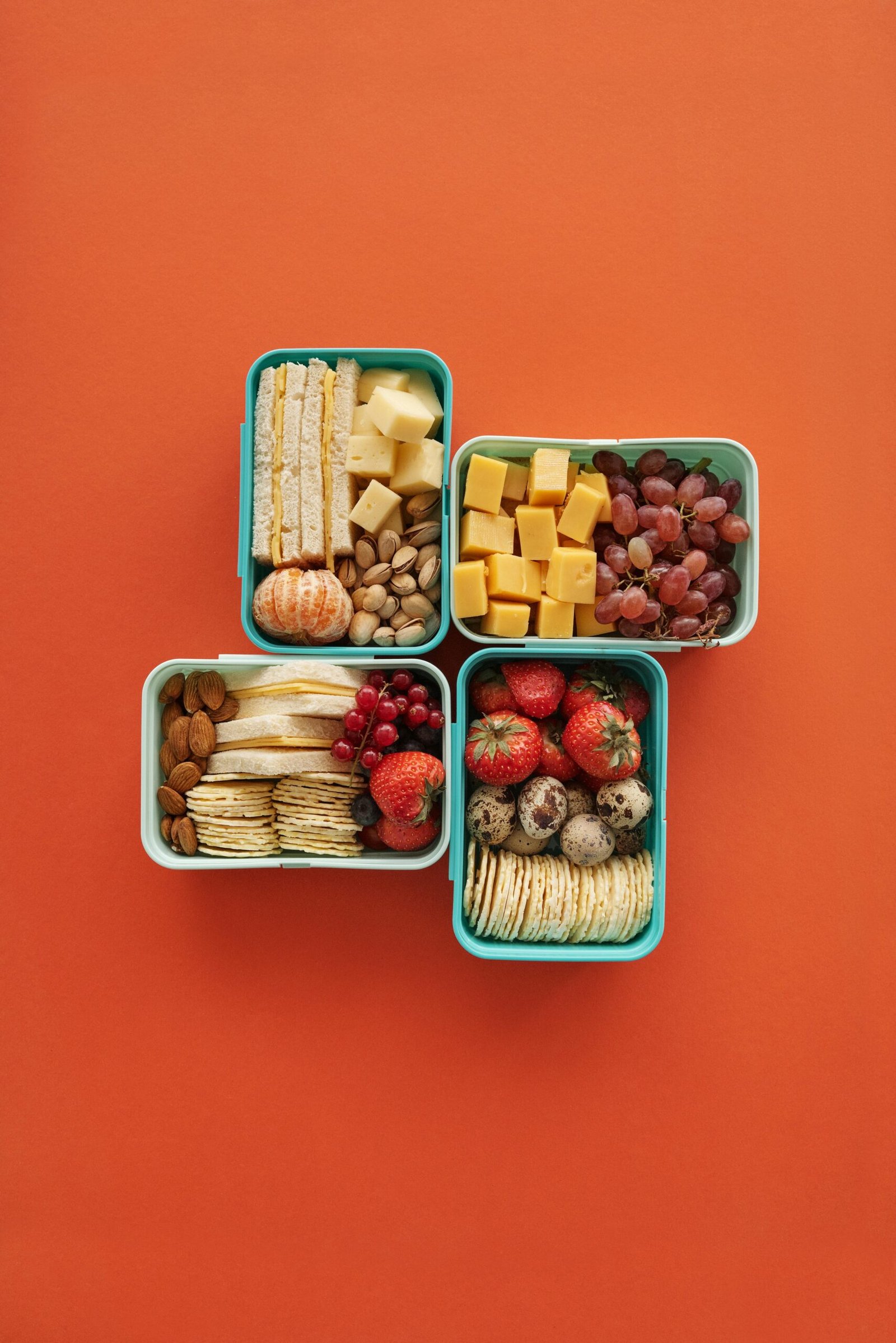Top view of four lunch boxes with fruits, cheese, and snacks on orange background.